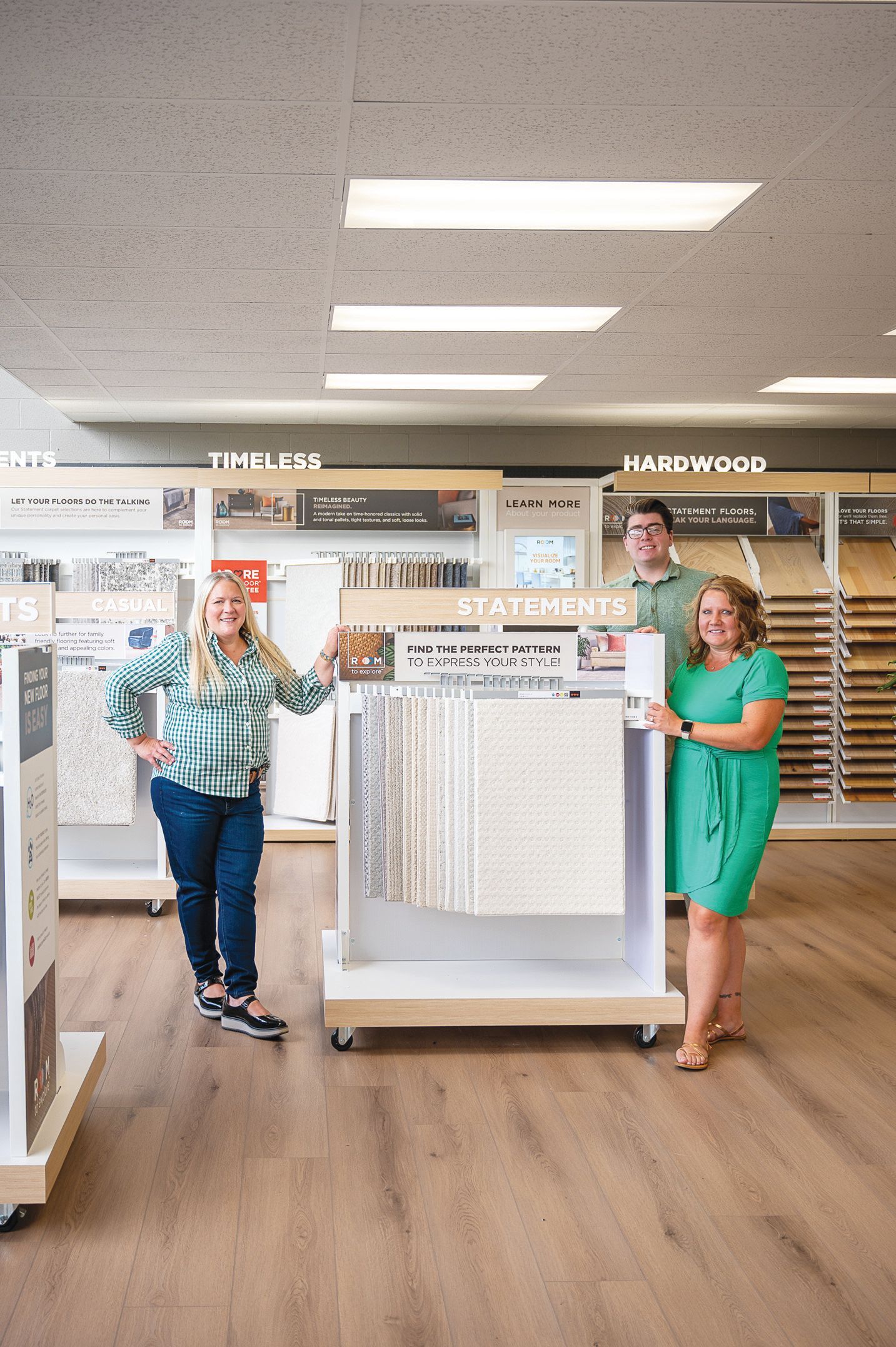 Two staff members and a man stand in a flooring showroom, showcasing a carpet sample display unit.
