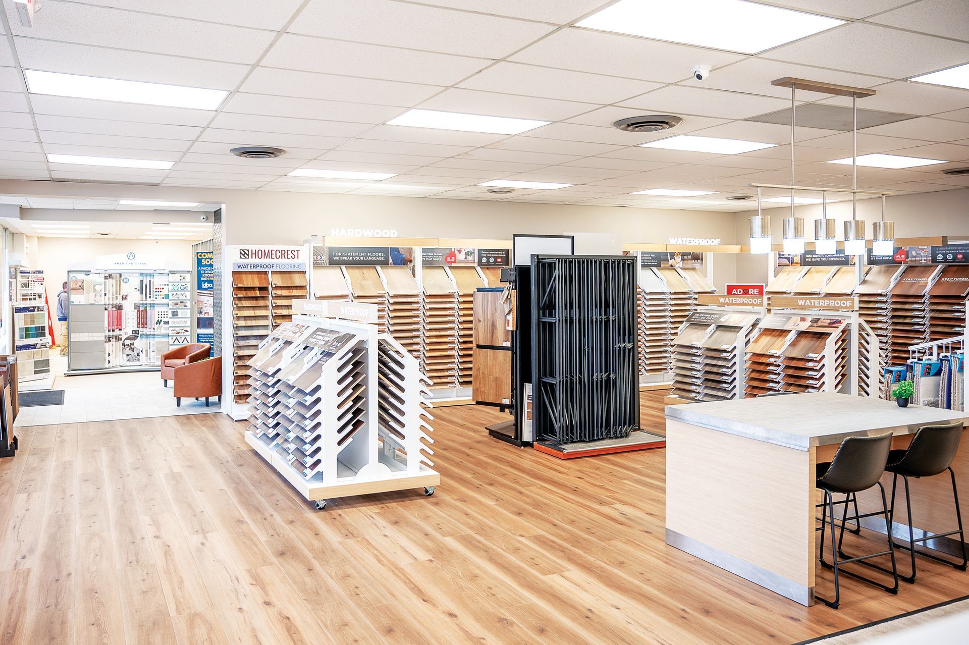 A bright, open flooring showroom featuring various wood plank displays, a central island with chairs, and tiled ceilings.