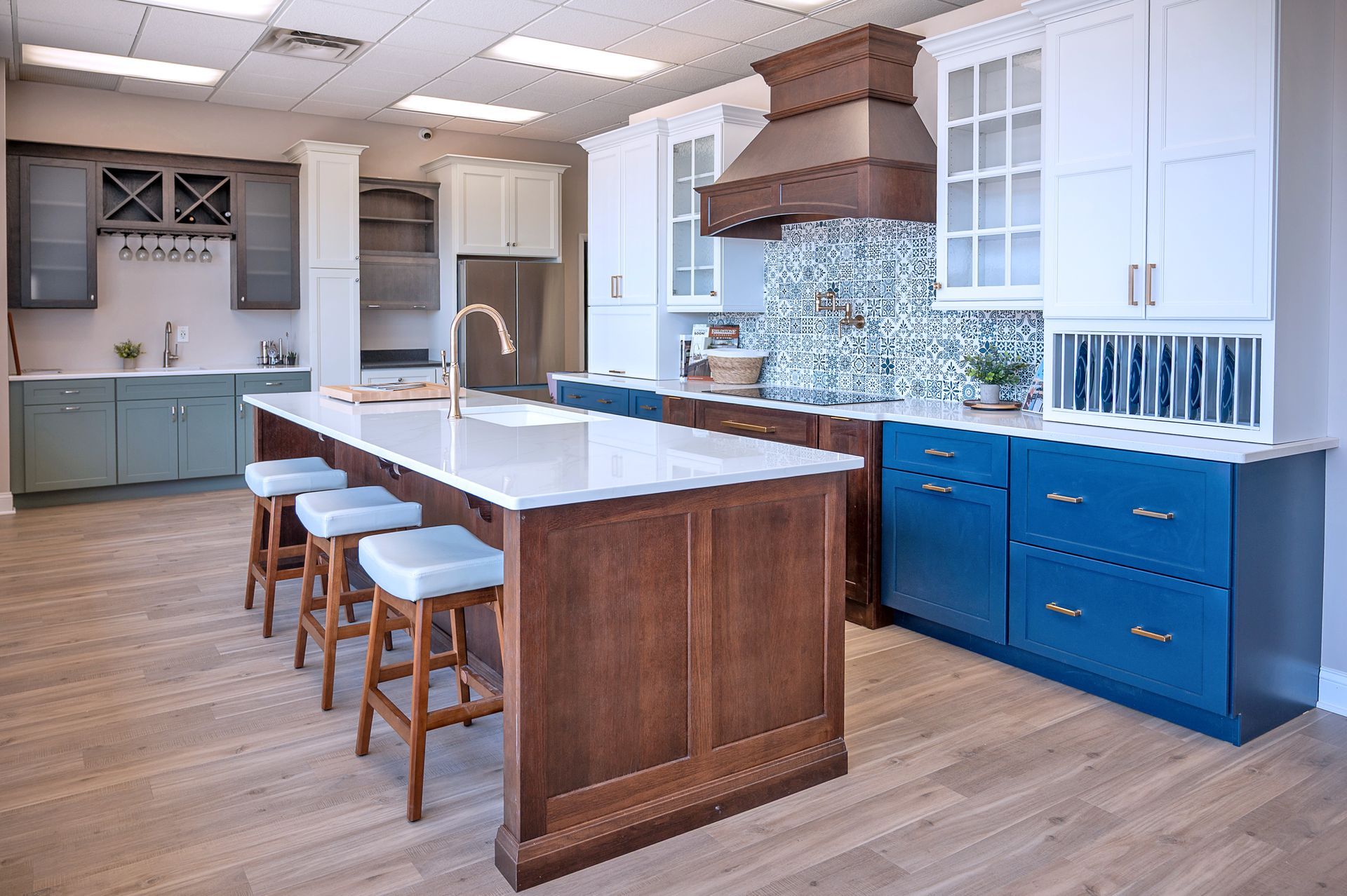 A kitchen showroom display featuring a wooden island with bar stools, blue lower cabinets, and a decorative tile backsplash.