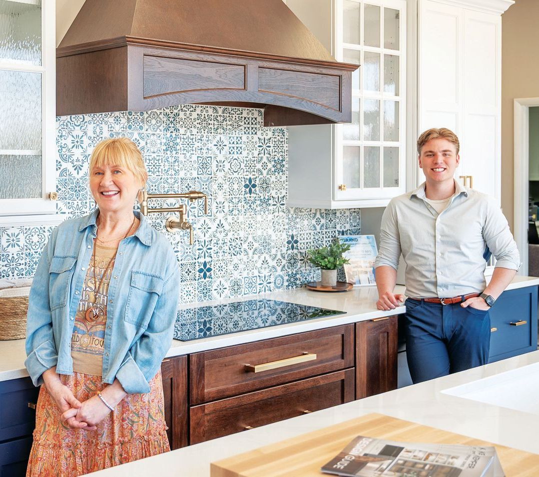 Two people stand in a kitchen with a wooden range hood, patterned blue tile backsplash, and dark cabinetry.