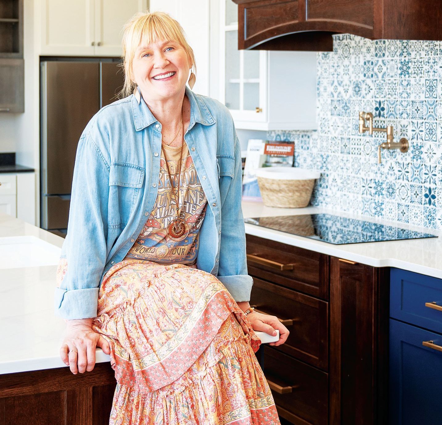A person with blonde hair smiles, wearing a denim shirt and patterned skirt, sitting on a kitchen counter near a stove.