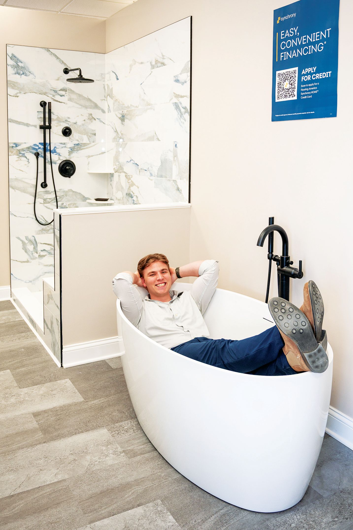 A person reclining in a white freestanding bathtub next to a marble-tiled shower display in a showroom.