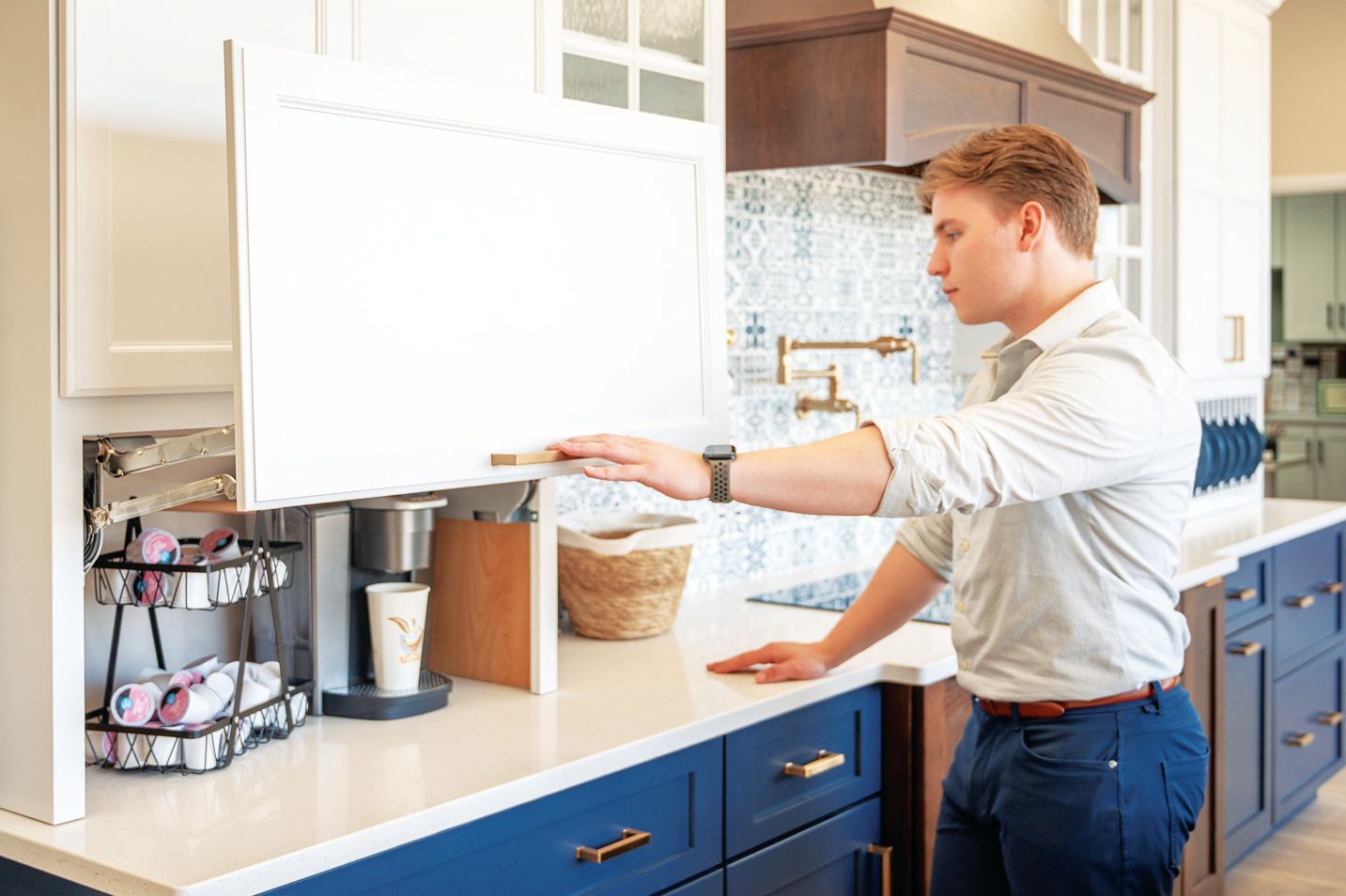 A person in a light shirt and blue pants lifts a white kitchen cabinet door to reveal a coffee station on the counter.