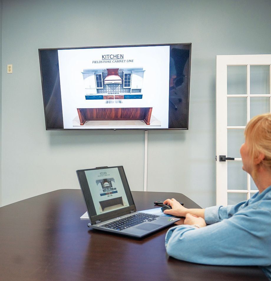 A person sits at a desk using a laptop that mirrors a kitchen design concept onto a wall-mounted television screen.