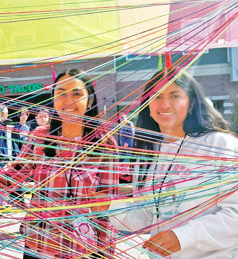 Two smiling individuals stand behind a dense, colorful web of crisscrossing yarn in an outdoor setting.