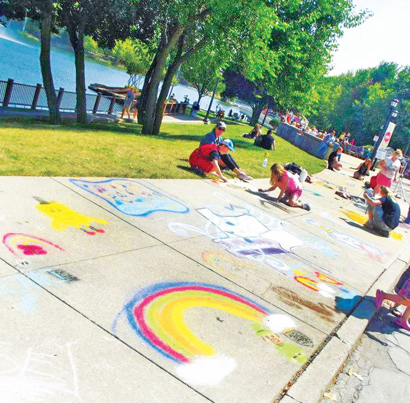 People draw colorful chalk art of rainbows, animals, and shapes on a park sidewalk near a lake on a sunny day.