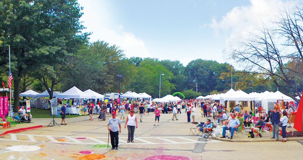 A sunny outdoor craft fair in a park with many white tents, people walking around, and colorful chalk art on the ground.