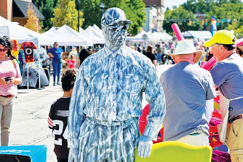 A street performer painted entirely in shades of blue stands in an outdoor market, surrounded by people and white tents.