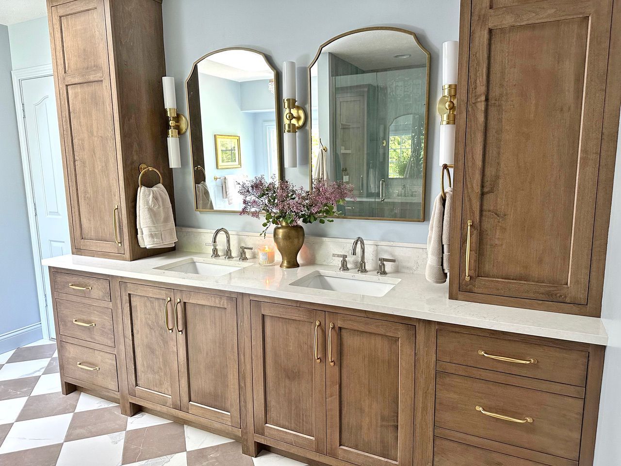 A wooden double vanity with white countertops, dual mirrors, and brass fixtures in a light blue bathroom.