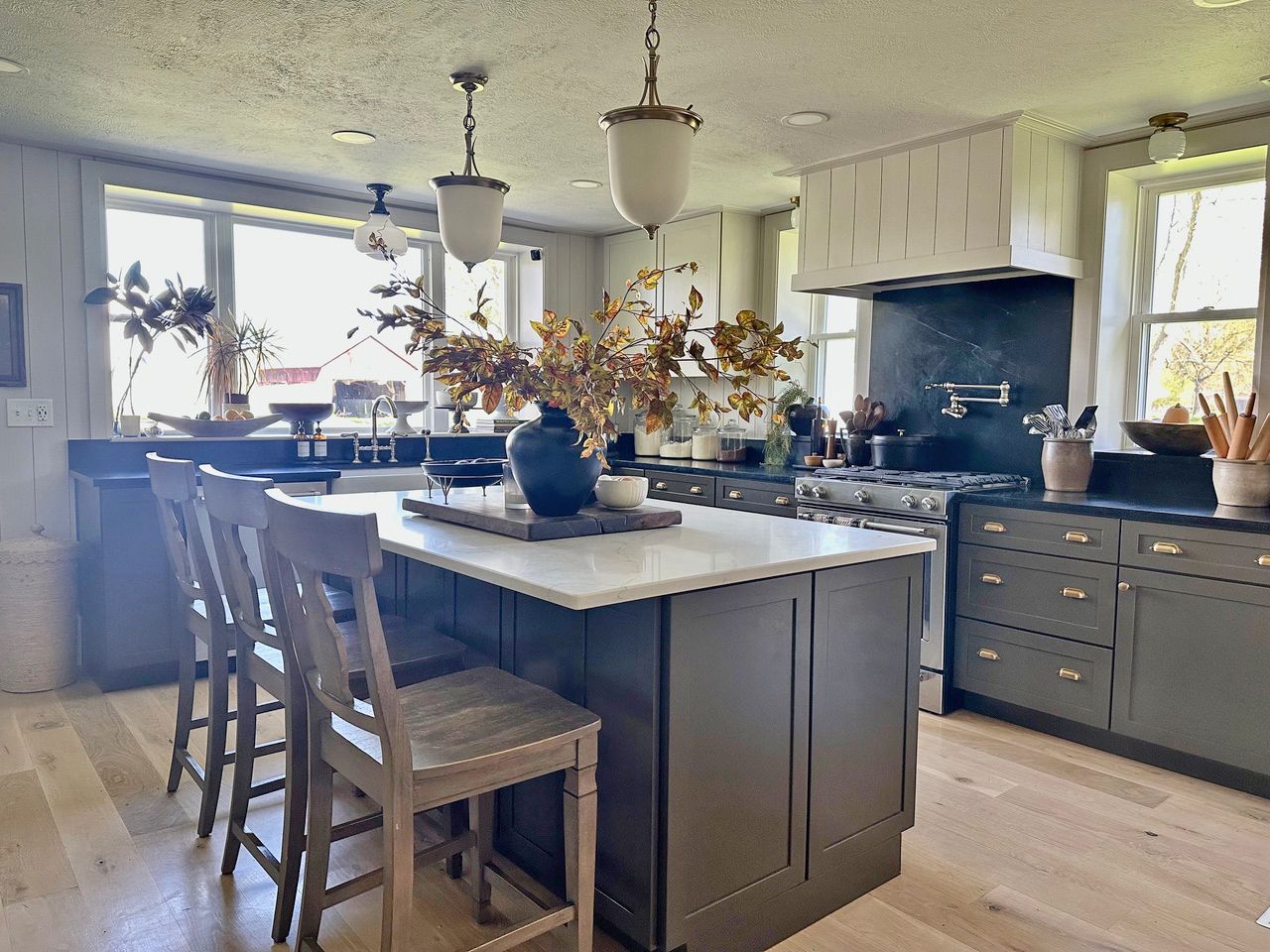 A bright kitchen features a large dark gray island with a white countertop, wooden bar stools, and a black range hood.