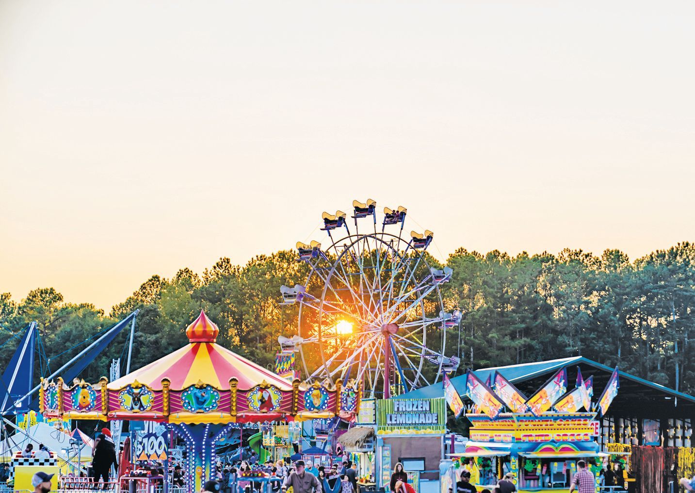 A colorful carousel and a Ferris wheel at a carnival during a sunset with trees in the background.