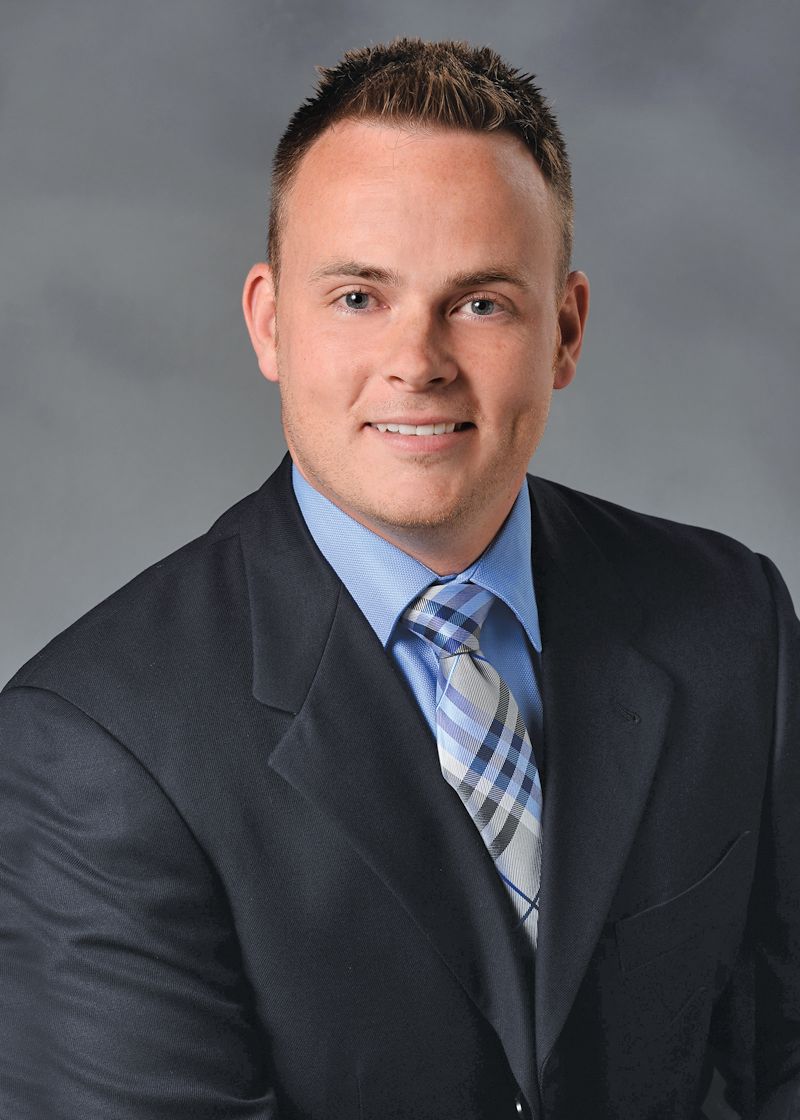 Headshot of a smiling person wearing a dark suit, light blue dress shirt, and a plaid tie against a gray background.