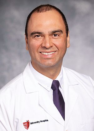 Headshot of a smiling medical professional in a white coat and purple tie against a blurred grey background.
