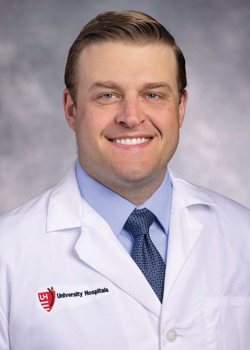 A person wearing a white lab coat with a University Hospitals logo, blue shirt, and patterned tie against a gray background.