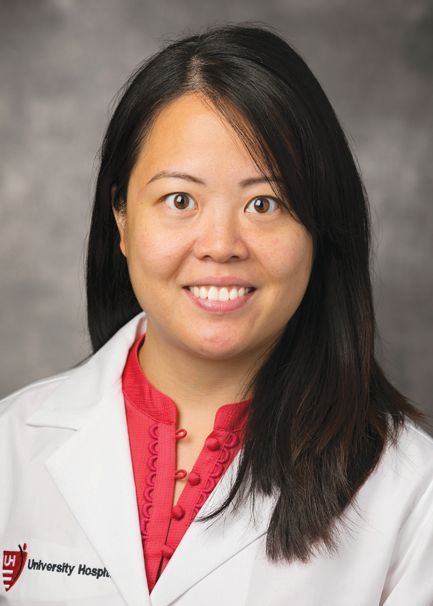 A professional headshot of a person in a medical coat with a red shirt, smiling against a gray backdrop.