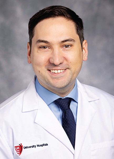 A healthcare professional in a white coat and blue tie smiles against a mottled gray background.