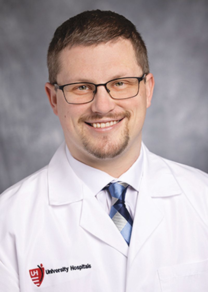 Professional headshot of a person wearing a white lab coat, glasses, and a blue tie, against a gray background.