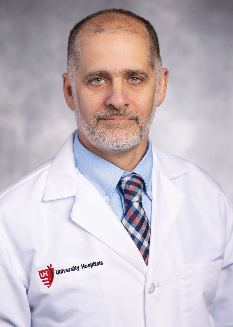 A professional headshot of a person wearing a white lab coat, blue dress shirt, and a plaid tie against a gray background.