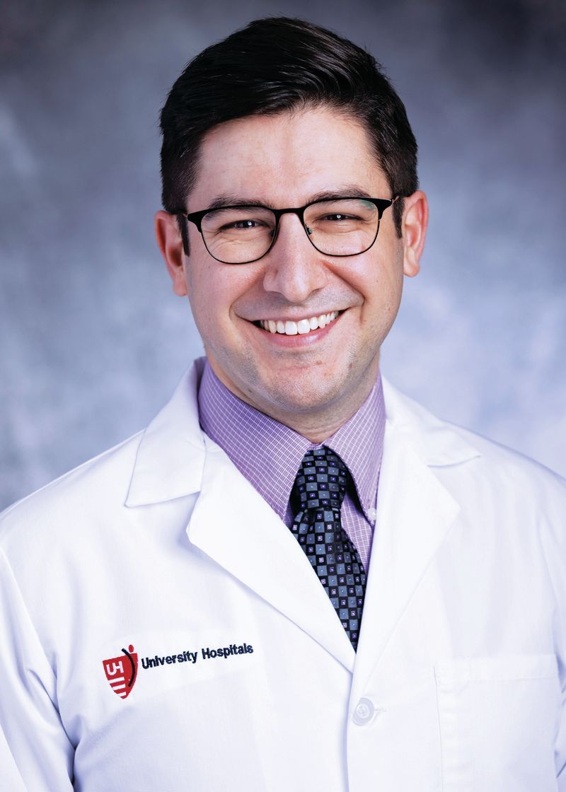 A smiling person wearing glasses, a purple dress shirt, a patterned tie, and a white lab coat with a hospital logo.