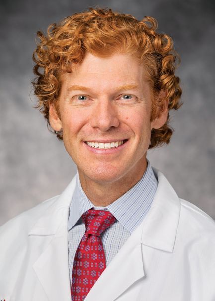 Smiling professional wearing a white lab coat, light blue striped shirt, and patterned red tie against a gray background.