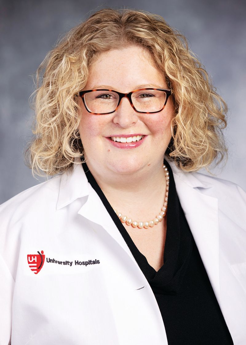 Smiling medical professional in a white coat with University Hospitals logo, wearing glasses and a pearl necklace.