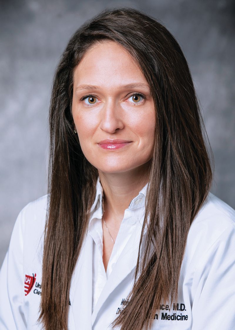 Headshot of a person in a white medical coat against a grey background.