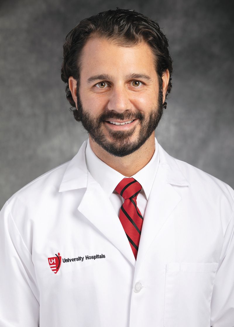 A healthcare professional in a white coat with a red tie and University Hospitals logo, smiling against a gray background.