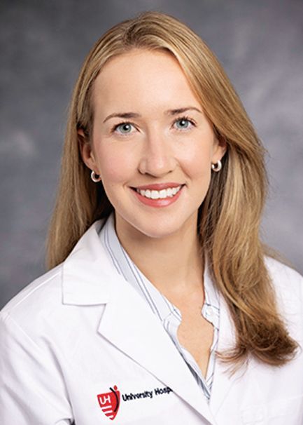 A smiling professional in a white medical coat and light blue shirt against a gray backdrop.