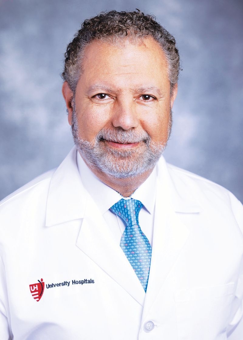A man in a white medical coat and blue tie with a University Hospitals logo, posing for a professional headshot.