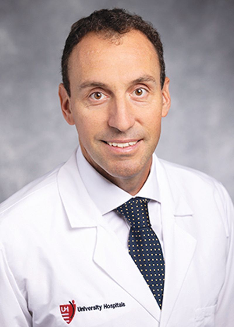 A professional headshot of a person in a white medical coat and a polka-dot tie standing against a grey background.