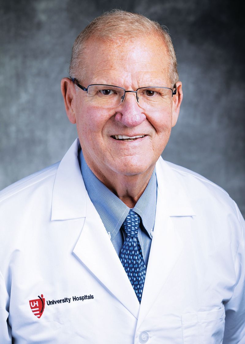 A doctor in a white coat with a blue shirt and patterned tie smiles against a neutral backdrop.
