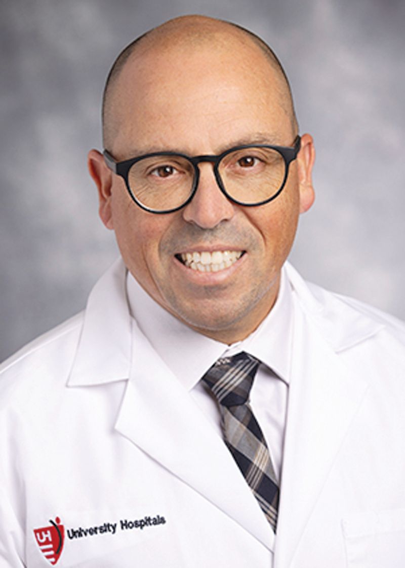 A smiling doctor in a white coat with a University Hospitals logo, wearing glasses and a tie against a gray background.