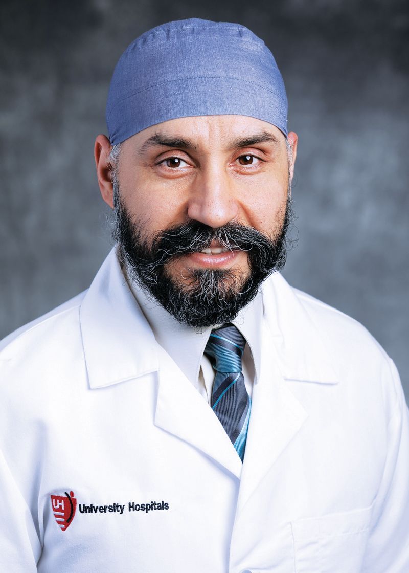 A medical professional in a white coat and blue surgical cap stands against a grey background, smiling.