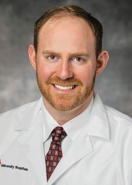Headshot of a smiling medical professional in a white coat and patterned tie against a grey backdrop.