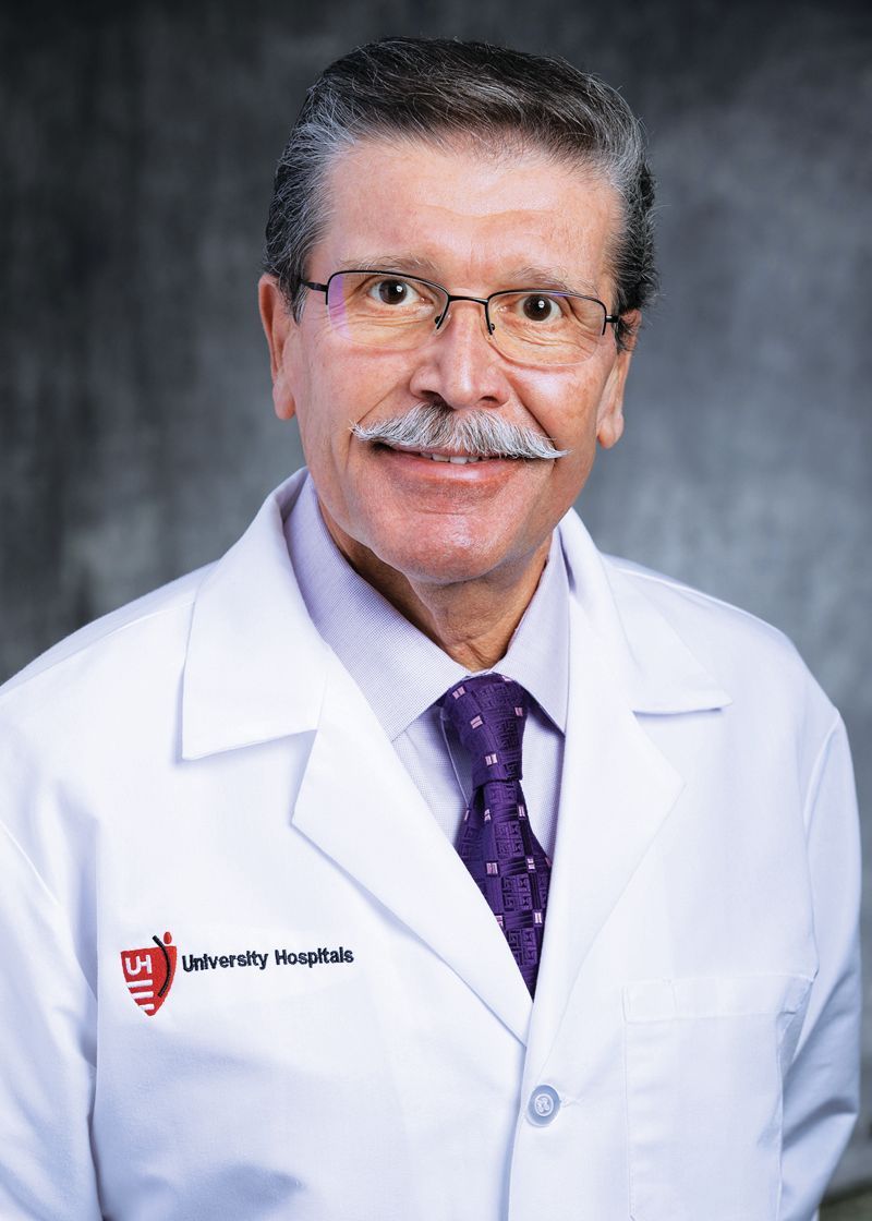 A man in a white lab coat with a University Hospitals logo and a purple tie against a grey background.