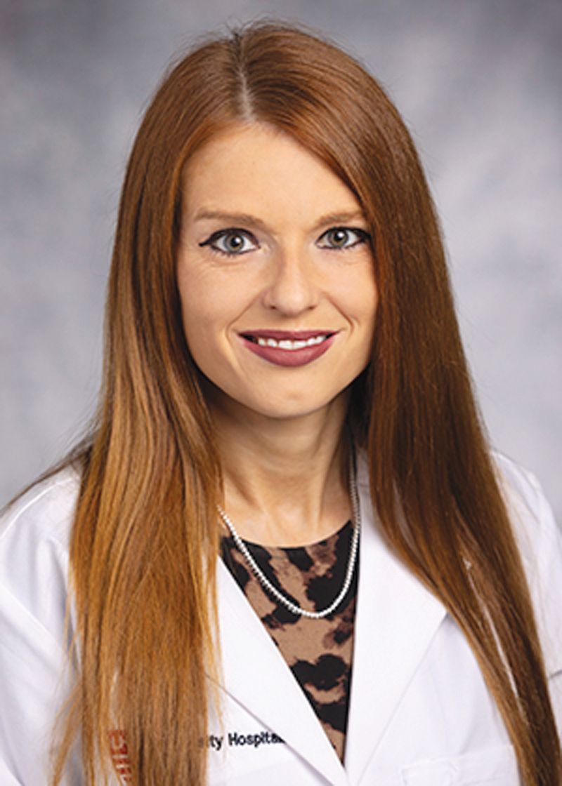 A professional headshot of a person with long reddish-brown hair wearing a white lab coat over a leopard-print top.