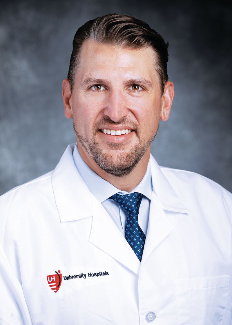 A person in a white medical coat and blue patterned tie smiles against a grey textured background.