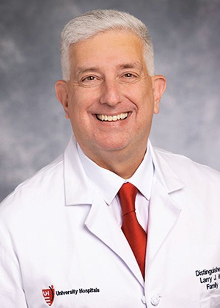 A person with short grey hair wearing a white lab coat and red tie, smiling against a mottled grey background.
