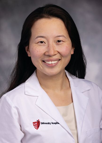 A smiling person wearing a white lab coat with a University Hospitals logo against a blurred, gray background.