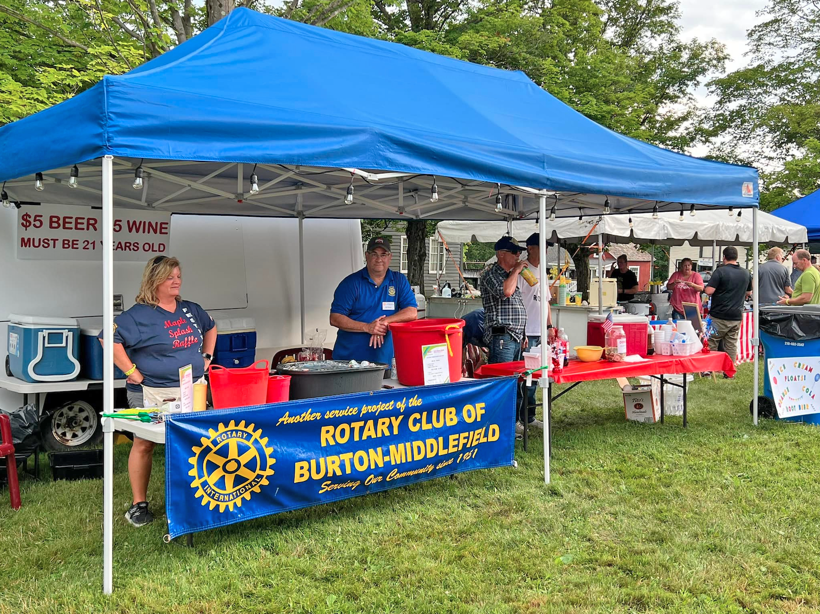 A blue tent housing a Burton-Middlefield Rotary Club booth at an outdoor event, with two people standing behind a table.