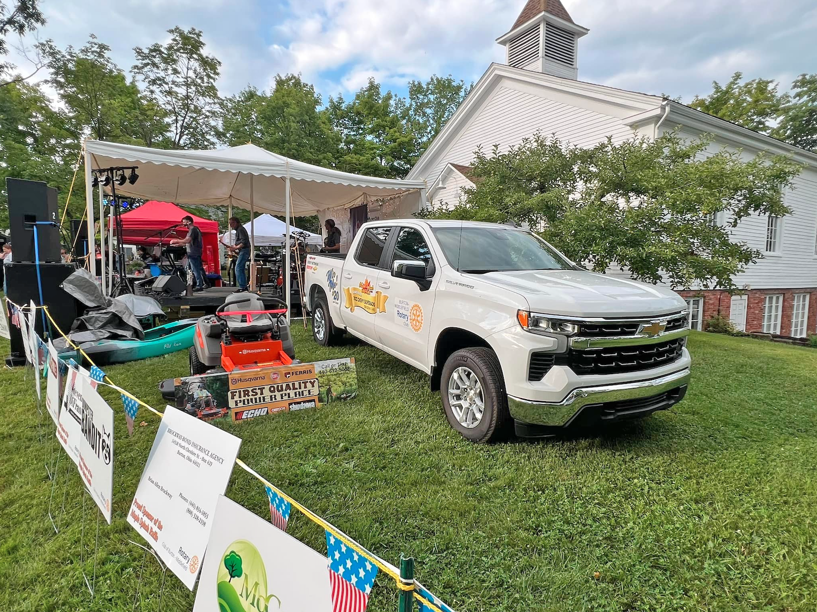 A white Chevrolet pickup truck sits on a lawn in front of a white church near a tent with equipment and a small ATV.