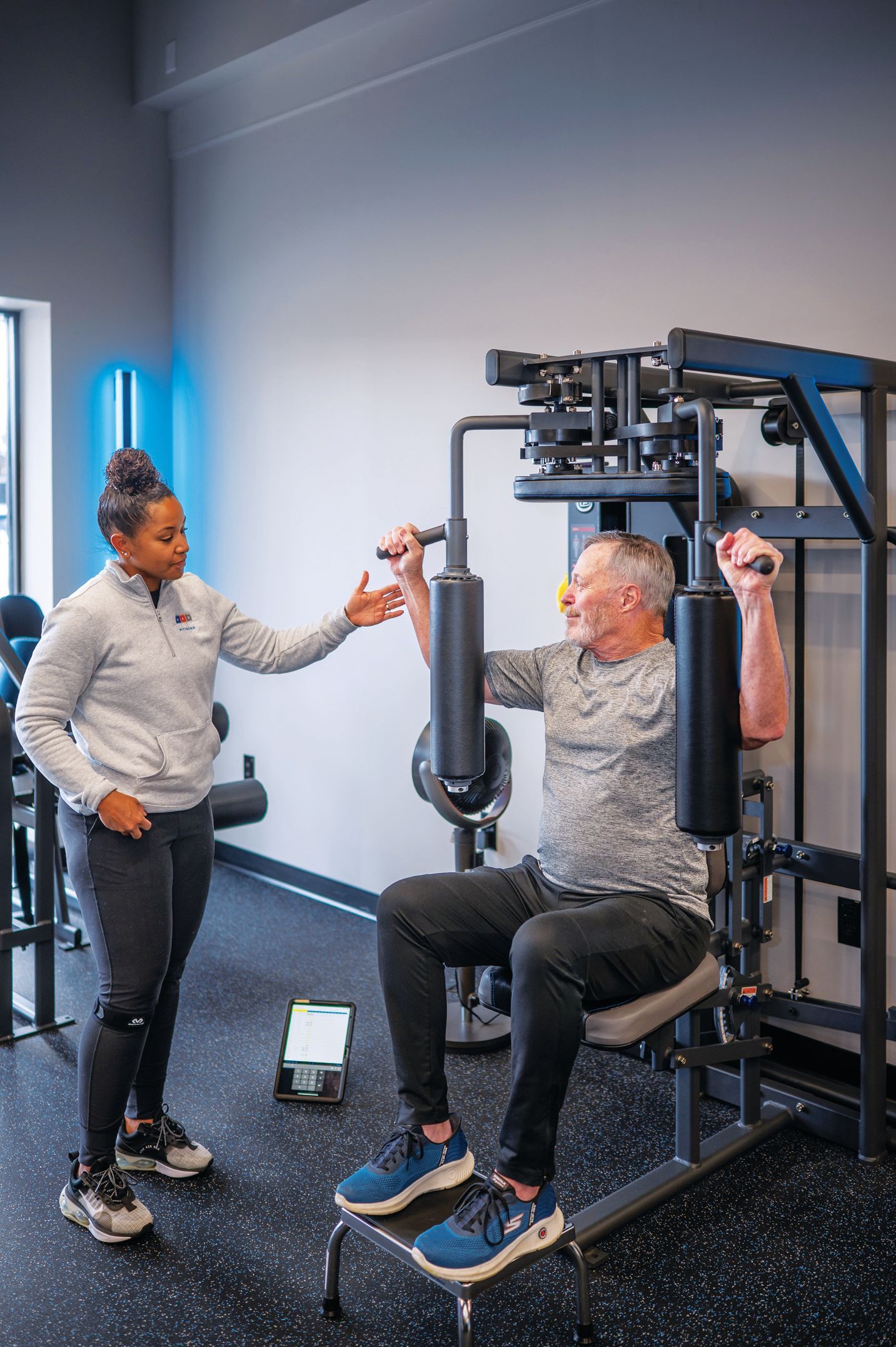 A fitness instructor observes an individual using a chest press machine in a gym.