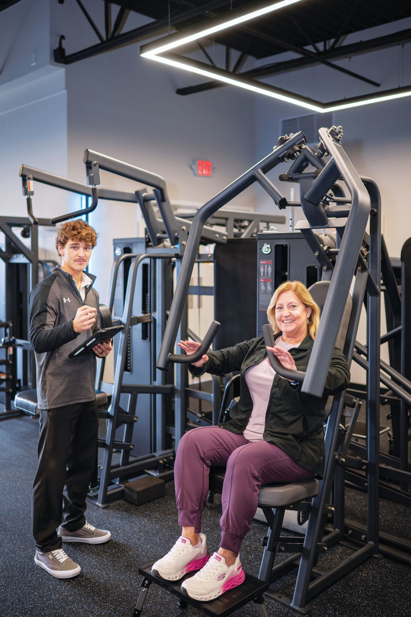 A fitness trainer stands with a tablet as a person uses a chest press exercise machine in a brightly lit gym.