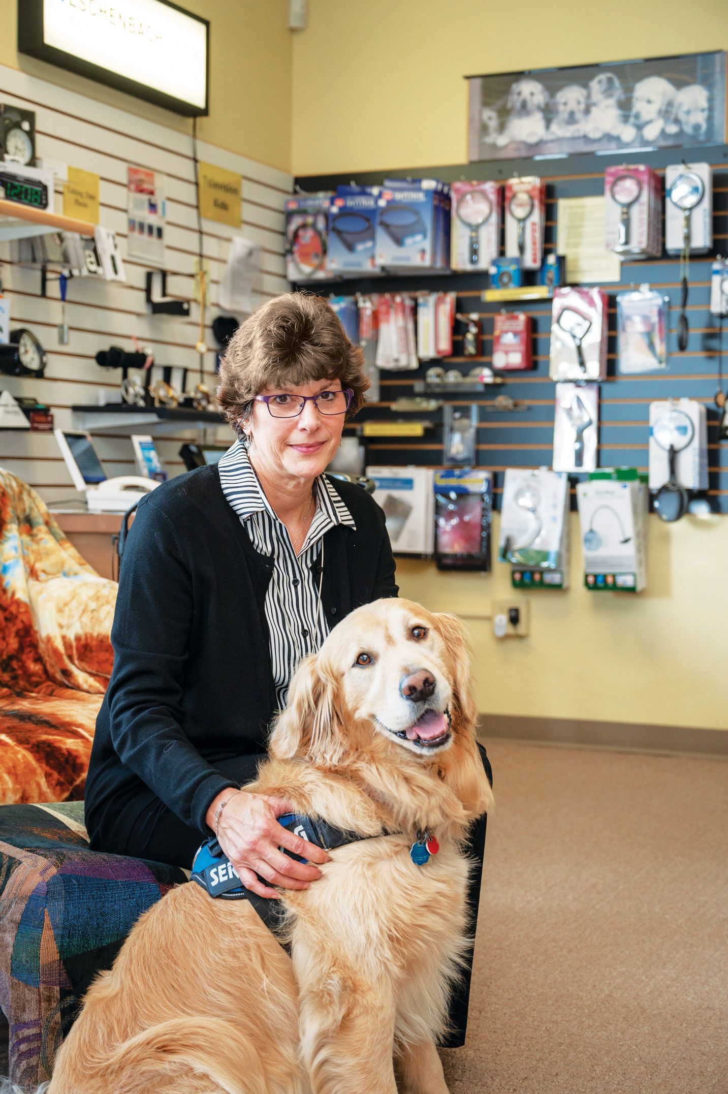 A woman sitting in a store with shelves of optical products, with a service dog resting at her side.