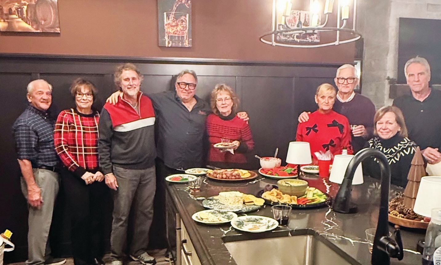 A group of nine people stand smiling together in a kitchen around a large island topped with food and serving platters.