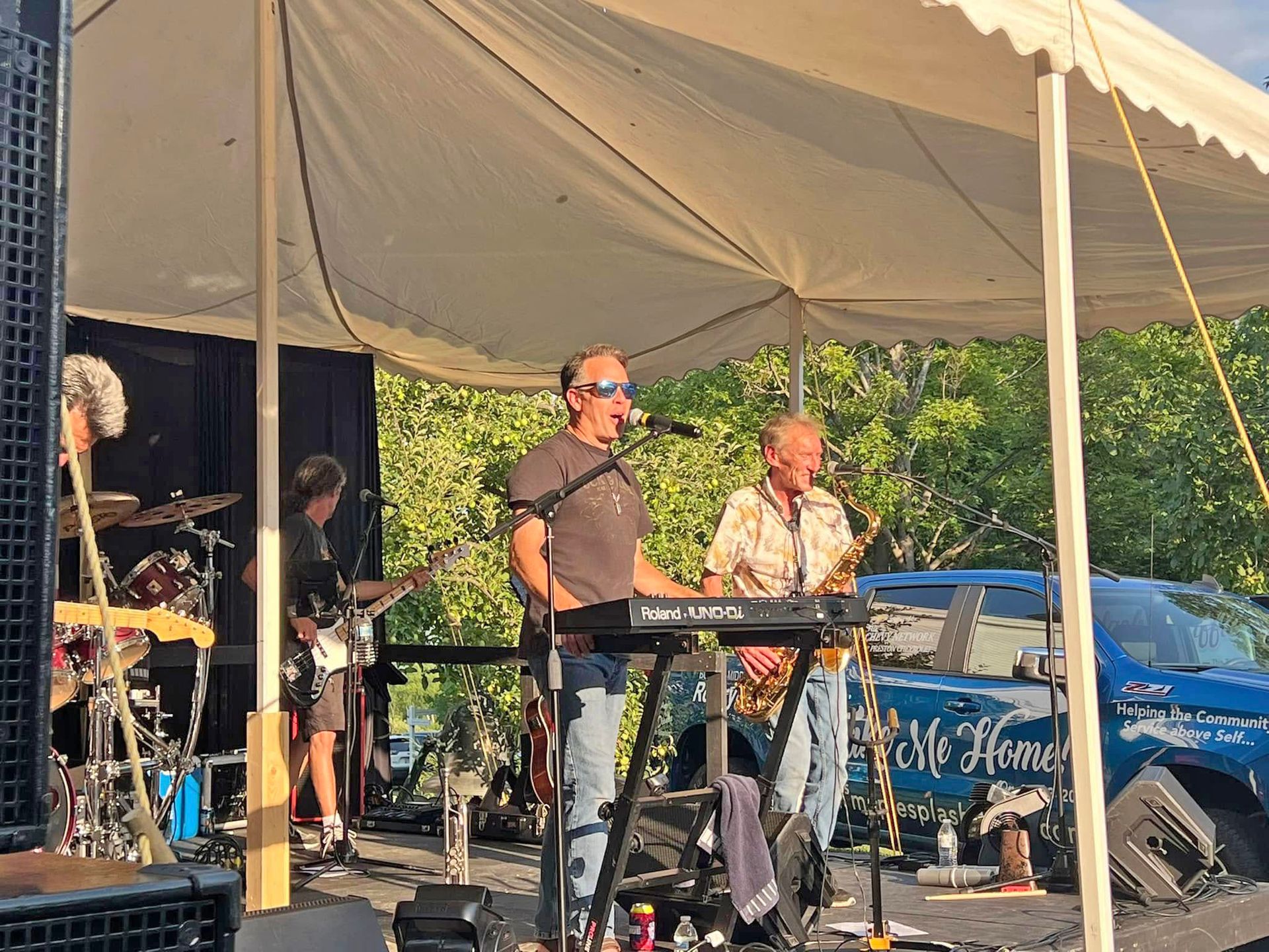 A live band performs under a white tent outdoors, featuring a keyboardist, a saxophonist, and a guitarist.