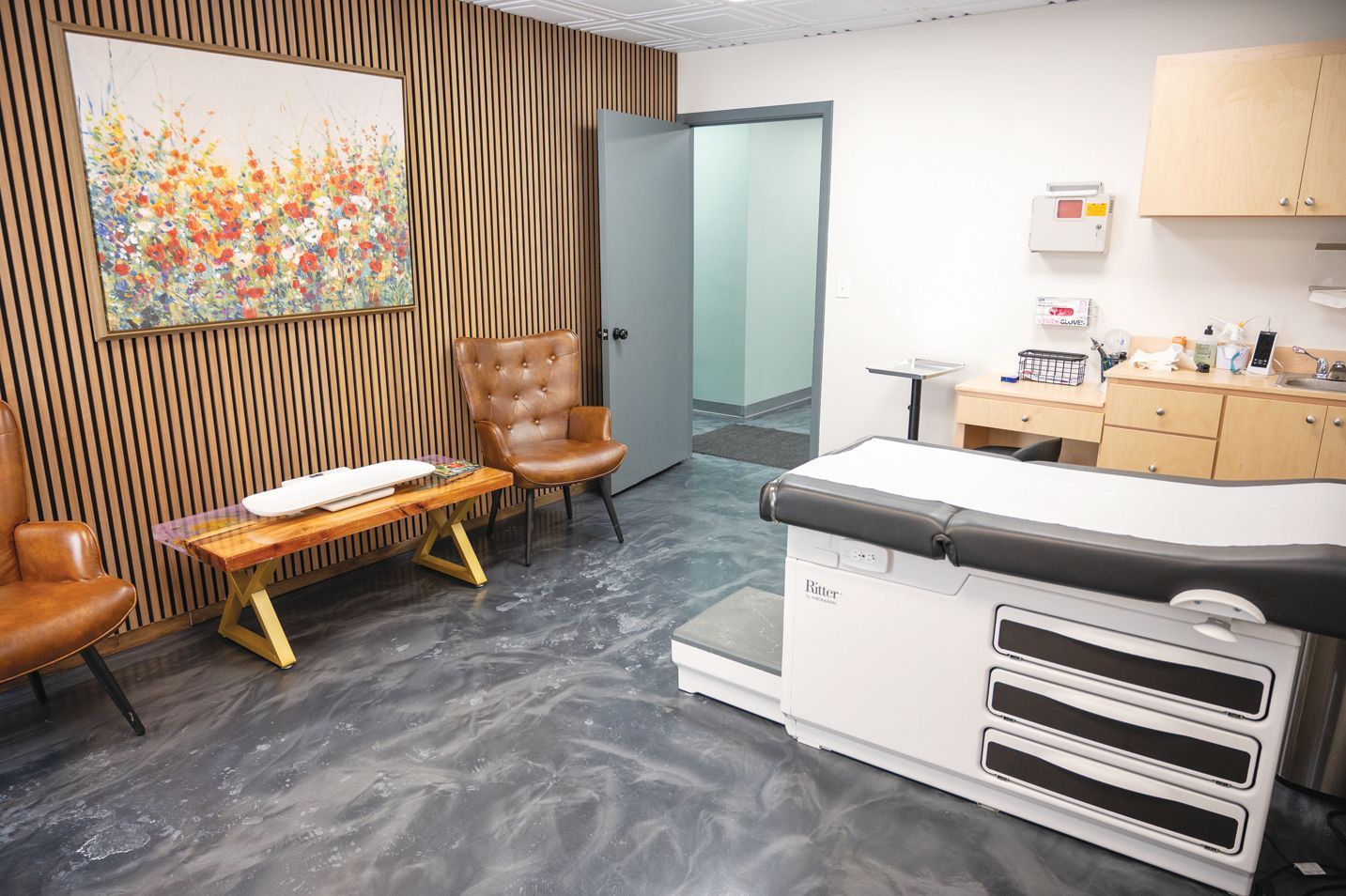 A medical exam room featuring an examination table, two leather armchairs, a wooden bench, and wall art.