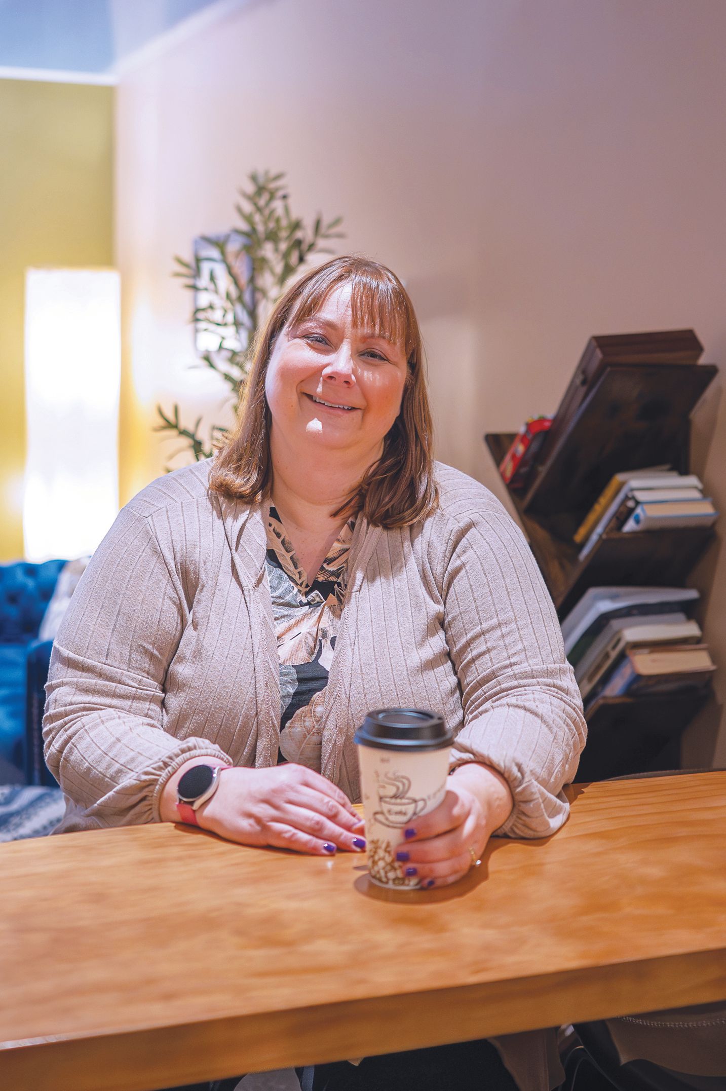 A person sitting at a wooden table holding a disposable coffee cup, smiling at the camera in a warm, indoor setting.