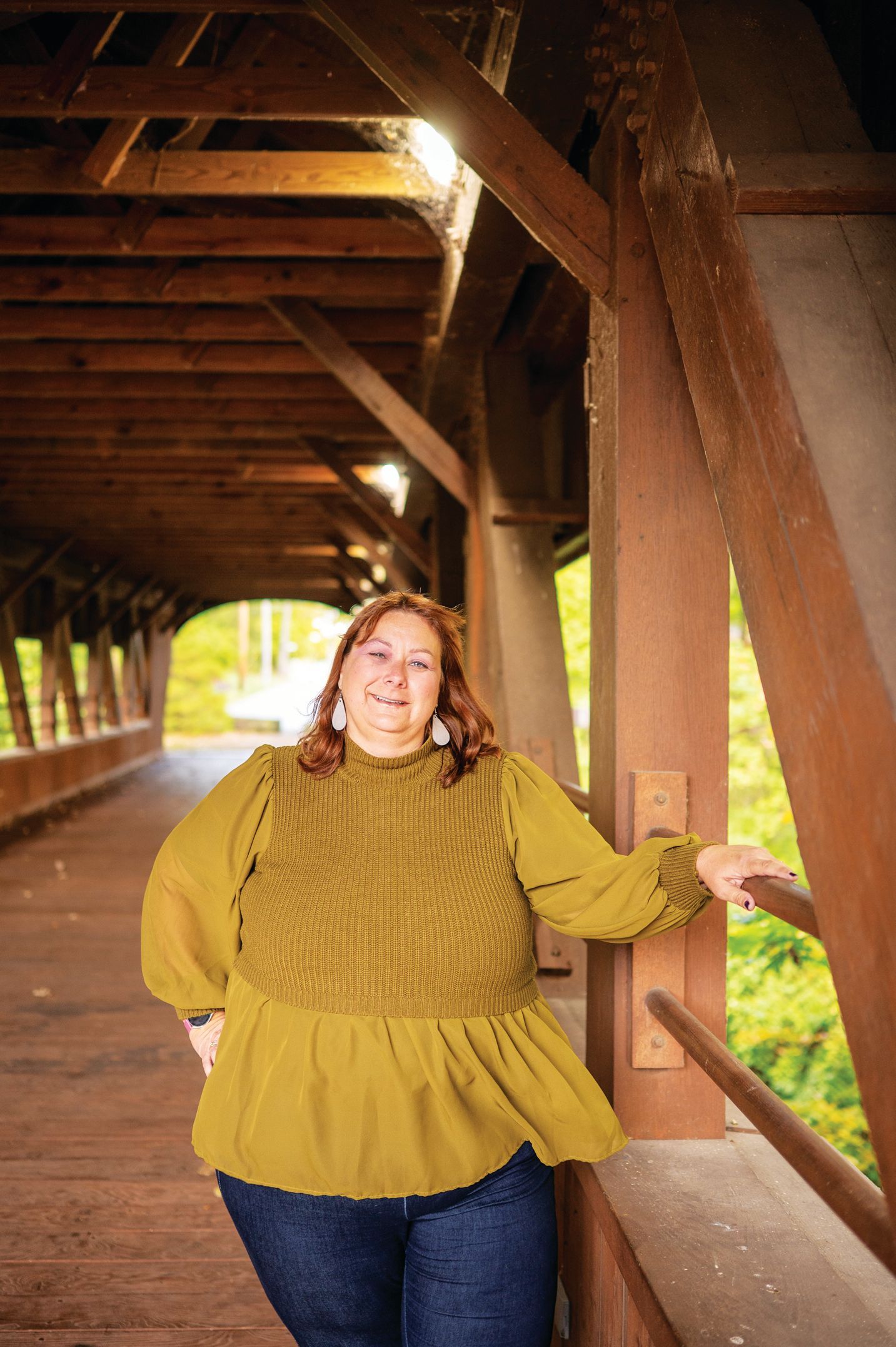 A person with red hair smiles, wearing a gold blouse and jeans, standing inside a wooden covered bridge.