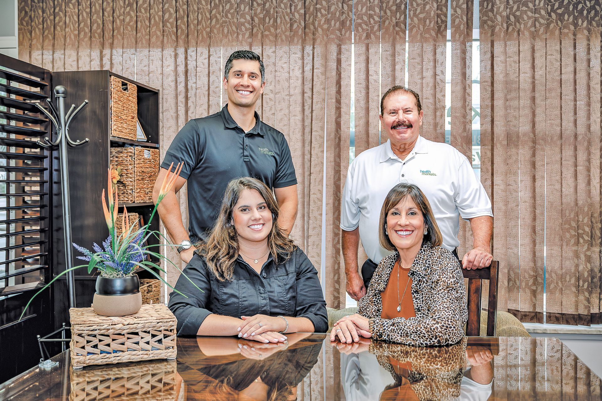 Four people posing for a professional portrait in an office setting with a desk, wood furniture, and blinds background.